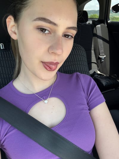 A fair-skinned young woman with brown eyes, light brown hair, and full lips is sitting in the passenger seat of a vehicle. She is wearing a purple ribbed top with a heart-shaped cutout at the chest. A silver necklace with a heart-shaped pendant is visible. A black seatbelt is fastened across her chest. The vehicle's interior includes dark grey patterned fabric on the seats. A glimpse of an exterior view, possibly trees or landscape, is visible through the vehicle's window. The location is not identifiable as the city of Levski, Bulgaria.