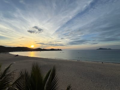 This image captures a serene sunset scene at a beach in Song Cau, Vietnam. The sun, a bright orb of yellow, is low on the horizon, casting a warm glow across the sky and reflecting on the calm sea. Wispy clouds, streaked with orange and yellow hues, paint the sky above. The beach itself is a expanse of soft, light-brown sand, with a gentle slope leading to the water. In the distance, lush green hills form a natural backdrop to the crescent-shaped bay. A few scattered palm fronds frame the foreground, adding a tropical touch to the scene. The water is a tranquil blue, with gentle waves lapping at the shore. A few small boats are visible in the bay, and a handful of people are scattered across the beach, some walking, others sitting, enjoying the peaceful atmosphere. One individual stands near the water's edge, while a couple is seated further down the beach. A red SUV is parked on the sand, its presence a subtle reminder of human activity in this otherwise natural landscape. The overall impression is one of tranquility and natural beauty, a perfect moment captured at the end of a day in Song Cau.