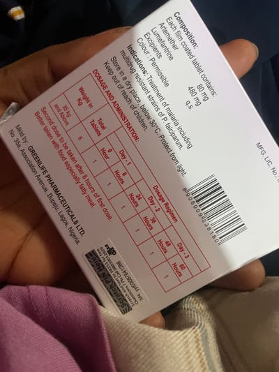 The image shows a close-up of a box of medication, likely an anti-malarial drug given the "Artemether" and "Lumefantrine" listed under "Composition". The box has dosage instructions and a table indicating the regimen based on weight and time. It also includes manufacturer information: "Mkt by: GREENLIFE PHARMACEUTICALS LTD." with an address in "Ilupeju, Lagos, Nigeria." The lighting suggests an indoor environment, and a hand is holding the box at an angle, obscuring parts of the text. The background is blurred, showing a dark fabric. There is a scratch-off panel for a PIN, indicating a security feature. The city of Iganmu is not directly mentioned or visible in the image.