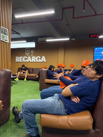 The image depicts a group of individuals, likely construction workers given their orange hard hats, resting on brown leather couches arranged on artificial turf. They are dressed in blue long-sleeved shirts and jeans. The setting appears to be an indoor waiting area or break room. One individual in the foreground is wearing work boots.

A digital clock on the wall shows the time as 10:21:10. Prominently displayed on the back wall is a large sign with the word "RECARGA" in white, block lettering, accompanied by a stylized logo of a battery and a plug. The environment is well-lit with overhead fluorescent lights. The overall mood seems relaxed, with individuals either resting, looking down at their phones, or conversing with each other. The location context is Villa El Salvador, Peru, which suggests this could be a facility or site related to work or services in that area. There is also a document with text and a QR code lying on a wooden surface in the foreground.