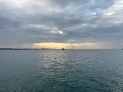 The image shows a body of water, likely the sea, under a cloudy sky. Beams of sunlight are breaking through the clouds, creating a dramatic effect. In the distance, a coastline is visible with what appears to be buildings or structures, as well as ships. The scene is in Türkiye.