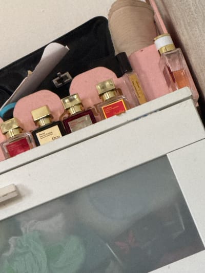 The image shows a collection of small perfume bottles, likely samples or travel sizes, arranged on a white shelf. The bottles have gold caps and some have labels indicating "Maison" and "Oud." Behind the bottles, a pink object, possibly a makeup bag or organizer, is visible. A black case is partially visible to the left of the pink object. The overall scene suggests a personal grooming or cosmetic setup. The background is a plain, light-colored wall. There is no visible indication of location within Аққыстау ауылдық округі, Kazakhstan.