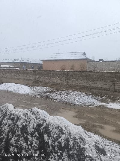 The scene depicts an outdoor environment in the city of Yakkabog, Uzbekistan, under snowy and overcast conditions, captured on January 16, 2026, at 16:26. The foreground features a large, dark pile of material, possibly dirt or gravel, heavily dusted with fresh snow on its surface and sides. Adjacent to this, the ground is a mix of wet earth, muddy patches, and scattered snow. A grey, modular brick or concrete block wall, approximately waist-height, extends horizontally across the mid-ground, serving as a boundary. Behind this wall, the ground is also partially covered in snow and wet patches.
In the background, a light brown, single-story building with a snow-dusted, dark corrugated metal gable roof is prominently positioned. A small, dark antenna or vent is visible on its roof. To the left and slightly further back, another single-story building with a flatter, snow-covered roof and at least two visible windows, one with a blue frame, is present. A second, lighter-colored brick or block wall, similar to the mid-ground wall, extends behind the brown building, forming another boundary. Bare tree branches are discernible above this wall. Multiple parallel power lines or communication wires stretch across the upper portion of the frame against a bright, uniformly grey overcast sky. No individuals, overt actions, or interactions are visible within the frame.