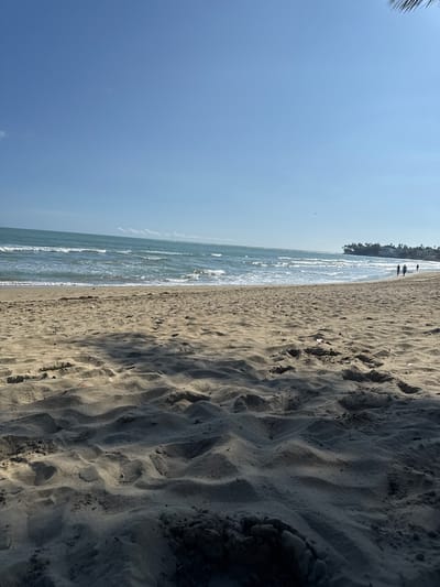 The image presents a beach scene in Cabarete, Dominican Republic. The shot is composed with the ocean and sky to the left and sandy beach to the right. There are a few distant figures walking along the shoreline near some buildings or foliage in the background. The sky is a clear blue. The beach sand shows variations in shading due to natural formations.