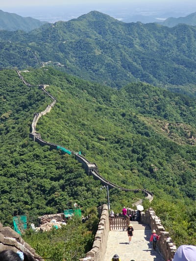 The image depicts a section of the Great Wall of China winding through a lush, mountainous landscape in Huairou District, China. The wall, constructed of stone and brick, is clearly visible, tracing the contours of the rolling green hills. Numerous tourists are present on the wall, walking along its path and descending a stone staircase in the foreground. Some are wearing hats and casual clothing, suggesting a warm, sunny day. The environment is dominated by dense green foliage covering the mountains, indicating a natural, possibly forested, setting. In the middle ground, there are some construction or repair activities underway, with scaffolding and green netting visible. The distant background shows a hazy expanse of sky and possibly a cityscape or more mountains, suggesting a vast, sprawling landscape. The perspective is from an elevated viewpoint, looking down upon the wall and the surrounding terrain.