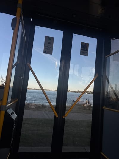 The image shows a view from inside what appears to be a bus, looking out through the doors towards the Swan River in Applecross, Australia. The doors have yellow grab rails across them and signage including "Caution: Mind the step, Doors open inwards" and "Did you tag off?". Beyond the doors, the river can be seen with a boat near the riverbank, along with some people. The environment consists of water, a grassy area on the riverbank, and a clear sky reflected in the glass.