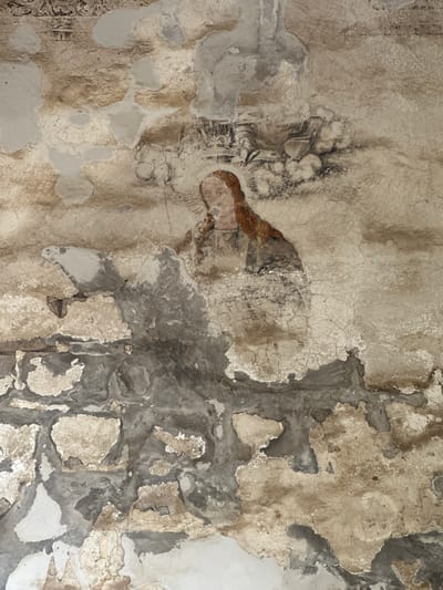 A faded mural depicts a figure with reddish-brown hair and a serene expression, rendered in earthy tones and dark outlines. The figure appears to be wearing a garment with a light-colored collar and possibly a darker overlay. Behind the figure, a shadowy, abstract form suggests a chariot or throne, complete with wheels and decorative elements, all depicted with similar muted colors. The mural is heavily damaged, with large sections of plaster flaking off, revealing the underlying stonework and rough texture of the wall. The damage creates an uneven surface, with lighter patches of new plaster repairs interspersed with the original weathered paint and crumbling substrate. The overall impression is of an ancient artwork in a state of significant decay, located within the historic structure of the San Miguel Arcángel Convent in Huejotzingo, Mexico.