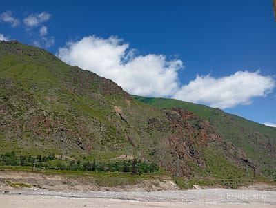 A wide, rocky riverbed is in the foreground, with a shallow, light-colored river flowing through it. Beyond the riverbed, a steep, green mountain rises, its slopes covered in grass and dotted with rocky outcrops. The upper parts of the mountain are a darker green, indicating denser vegetation. A few power lines and pylons are visible on the lower slopes, along with a line of trees and some structures that suggest a settlement or road. The sky is a bright blue, with scattered white clouds. The scene is located near Tyrnyauz, Russia, as indicated by the date stamp "01.07.2026 10:47" in the lower right corner, which is a future date.