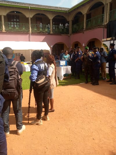 The image shows a group of people gathered in the courtyard of a building in Beni, Democratic Republic of Congo. The building appears to be a school, with a second-story balcony overlooking the courtyard. The ground is dirt, and there is a white tent in the foreground. In the center of the courtyard, a group of people are standing around a blue box, which is being held by a person in a green uniform. There are several people with cameras and video equipment. The majority of people are wearing casual clothes, with some men wearing uniforms. One woman is wearing a blue shirt with a "PRESS" vest. It seems there is a security detail present, including a man wearing a green uniform. The scene is focused on the group of people gathered around the blue box. The image is taken from a low angle, looking up at the people gathered around the box.