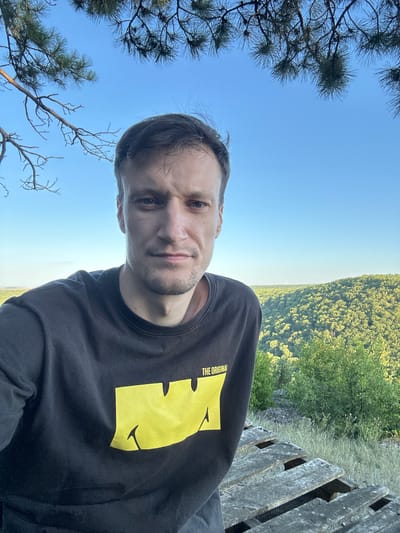 A man takes a selfie outdoors in the city of Pryvorottia Druhe, Ukraine. The man is wearing a black t-shirt with a yellow graphic. He is in the foreground, with a lush, green forest and a clear blue sky in the background. There are also some trees branches at the top of the image. The man is looking directly at the camera. A wooden pallet is visible on the right side of the image.