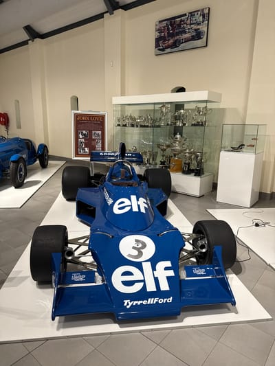 This is an indoor shot of a Formula 1 race car, specifically the Tyrrell 006, exhibited in a museum or similar display setting. The car is predominantly blue with white "Elf" logos and the number "3" prominently displayed. "TyrrellFord" branding is also visible. The car is mounted on a white platform.

Behind the race car are glass display cases containing numerous silver trophies and racing memorabilia. To the left of the race car, there's a framed display about John Love, featuring photographs and text related to his racing career. A framed photograph is visible on the wall above the trophy cases. The location is the Franschhoek Motor Museum in the Stellenbosch Local Municipality, South Africa.