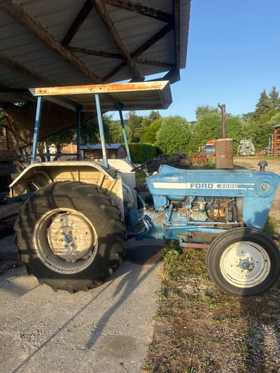 This is a medium shot, eye-level, outdoor, daytime photograph of a blue and white Ford 3900 tractor parked under a rustic, covered structure. The structure has a corrugated metal roof supported by wooden beams and a rusty metal frame. The tractor is positioned on a concrete surface, with dry grass and dirt surrounding it. The tractor's engine is visible, and it has large, worn tires. In the background, there are trees, a clear blue sky, and other structures including a shed and what appears to be construction equipment. The sun casts long shadows from the tractor and the supporting structure. The image appears to be taken in Charchilla, France, given the context of the location being provided. There are no people visible in the frame.