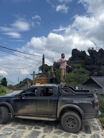 The image shows a man standing on the roof of a black "RAPTOR" pickup truck in Tả Van, Vietnam. He is fair-skinned, with a short beard, wearing a pink shirt and tan pants, holding an object in each hand, and looking towards the right of the frame. Behind him is a rocky outcrop with a sign on top, accessible by a series of stairs. Below this is a building with a brown roof. The truck is parked next to a sign that says "ROCK GARDEN HOMESTAY - COFFEE - BBQ TUYEN NHAN VIEN". Power lines are visible overhead. The sky is blue with scattered clouds. The ground is paved with stone. The truck appears to be covered in mud.