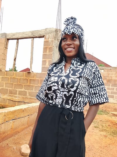 A young woman stands in front of a partially constructed brick building in Barikin Sabon Gidan Kanat, Nigeria. She wears a black and white patterned top and matching headwrap, paired with black pants. Her right hand is in her pocket, and she is smiling and looking to her left. The building behind her has open window frames with wooden supports. In the background, there is a glimpse of a red-roofed structure and an overcast sky. The ground is reddish-brown dirt.