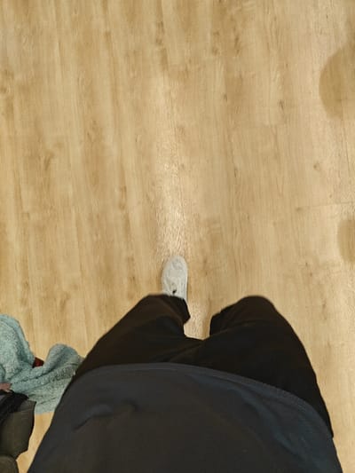A first-person perspective shows a person standing on a light wooden floor. The person is wearing black pants and white sneakers. A light blue towel is visible to the left, on top of some dark-colored items, possibly a bag. The focus is on the lower body and the immediate surroundings, indicating an informal and personal setting.