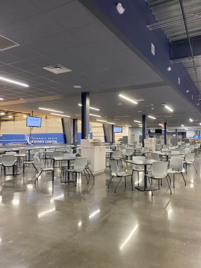 The image depicts an interior view of the Virginia Beach Sports Center, located in Virginia Beach, United States of America. The scene is dominated by rows of empty tables and chairs, set on a polished concrete floor that reflects the overhead lighting. The tables are square and dark-colored, with matching dark-colored chairs featuring thin metal legs.
In the background, a large blue wall with white lettering reads "VIRGINIA BEACH SPORTS CENTER". Above this wall, a screen displays "CODE OF CONDUCT". Further in the background, a sports netting structure is visible, suggesting the presence of courts or fields. The ceiling is a dark gray drop ceiling with numerous fluorescent light fixtures. There are also several dark-colored support columns spaced throughout the area. The overall environment appears clean and well-maintained, designed for a public facility. There are no people visible in the image.