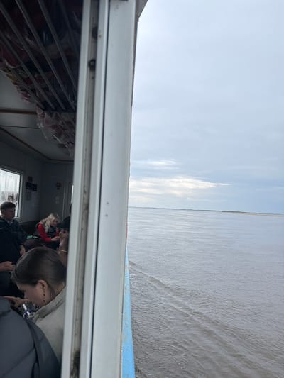 The image captures a scene from a boat or ferry, looking outwards over a vast body of water while simultaneously showing a glimpse of the passenger cabin.

In the foreground, occupying the left and bottom portions of the frame, is the interior of the boat's cabin, viewed through an open doorway or large window frame. Several passengers are visible, mostly seated. A woman with blonde hair, wearing a red and black striped top, is clearly seen seated and looking towards the front of the boat. Other passengers, identifiable by their heads and shoulders, wear casual clothing such as jackets and hoodies. Along the left wall of the cabin, rows of red safety equipment, likely life vests, are neatly stored in clear plastic bags within metal racks. Further inside, cabin features like lights and unreadable signs are discernible on the wall.

Beyond the boat's interior, the external view is dominated by a wide expanse of calm, grey-blue water, suggesting a large river or lake. The water surface shows subtle ripples and small waves, indicating movement. A thin strip of land or shoreline is visible on the distant horizon, emphasizing the breadth of the waterway. Above the water, the sky is partly cloudy, with soft white clouds interspersed with patches of light blue. A section of the boat's blue and white exterior railing is visible along the lower edge of the outer view.

The scene depicts a peaceful daytime journey. The lighting suggests a bright, possibly overcast day, either in the morning or afternoon. No visible text is clearly legible within the image.