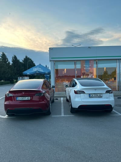 The scene depicts an asphalt parking lot in Momkovo, Bulgaria, at dusk or early evening, facing a commercial building. Two Tesla Model Y vehicles, both with Bulgarian license plates, are parked in marked spaces. The car on the left is dark red with license plate "EA 8899 AB". The car on the right is white with license plate "EA 3306 AE" and features a "DUAL MOTOR" badge.
The commercial building on the right has a light-colored facade with a green horizontal stripe above large glass windows that reflect the sky and surrounding elements. A poster visible within the window displays Cyrillic text, likely "Време за нови вкусове" (Time for new tastes), alongside images of various cold beverages. Two surveillance cameras are mounted on the building's exterior.
To the left of the building, dark green coniferous trees are visible, behind which a light-colored, small building or utility structure is partially seen. A large blue outdoor umbrella, branded with "DEVIN Minerals & Vitamins" in white text, stands between the trees and the commercial building. A double-armed streetlight is positioned above the umbrella.
The sky exhibits a gradient, transitioning from a pale yellow-orange hue near the horizon on the left to a lighter blue further up and to the right, with scattered light clouds. A small, dark object, possibly a bird, is visible in the upper left quadrant of the sky. No individuals are present in the image.