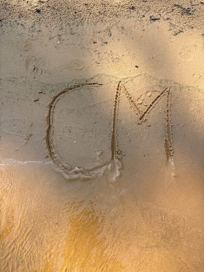 The image shows the letters "CM" written in the sand on a beach, with the water gently lapping at the lower portion of the image. The sand is a light brown color, with some darker areas and debris visible. The letters are formed by clear indentations in the sand, with a slight ripple effect from the water. The overall scene is one of a natural outdoor environment.