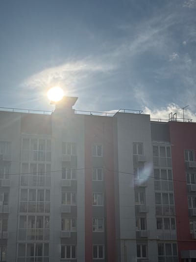 The image captures a modern apartment building in Volokolamsk, Russia, under a bright, partly cloudy sky. The sun is high and bright, creating lens flares that streak across the frame and partially obscure the upper portion of the building. The building itself is constructed with sections of light gray and a vibrant pinkish-red. Numerous windows and balconies are visible, many with white railings. Air conditioning units are mounted below some of the windows. A telecommunications antenna is mounted on the roof of the building on the right side. The scene appears to be during daylight hours, likely morning or afternoon, given the sun's position and intensity. There are no people or vehicles immediately visible in the frame. The overall impression is of a residential area bathed in strong sunlight.