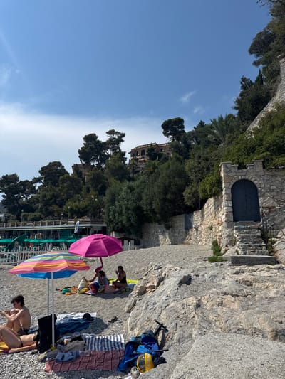 The image depicts a scene on a pebble beach in Finale Ligure, Italy, with a bright blue sky overhead. In the foreground, several people are relaxing on towels under colorful beach umbrellas. Two umbrellas, one striped and one magenta, are prominent. A young man is seated on a towel on the left, reading a book, and a woman is sunbathing nearby. Further back, under the magenta umbrella, a family appears to be having a picnic.

The beach is made of small pebbles, and large rocks are visible on the right side of the frame. Behind the beach, a stone wall runs along the hillside, topped with lush green trees and vegetation. A stone structure with a dark, arched double door and a stone staircase leading up to it is situated on the right. Above the trees, a multi-story building with terracotta roofs is visible on the hillside. In the background, a row of green beach umbrellas and a structure with a white railing suggest a beach club or restaurant. The overall impression is a sunny day at a picturesque Italian beach.