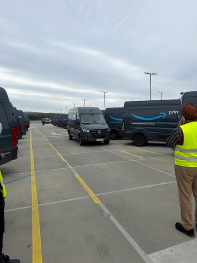 A long row of dark gray Amazon Prime delivery vans is parked in a large outdoor parking lot. The parking lot is marked with yellow and white lines. Some individuals wearing yellow vests stand near the vans, possibly inspecting or preparing them for deliveries. The sky is overcast, suggesting a cloudy day. The vans are predominantly of the Mercedes-Benz Sprinter type. The license plate of one of the vans is visible, showing it is registered in Germany, specifically "MZ." The location is likely in Friedrichsdorf, Germany.