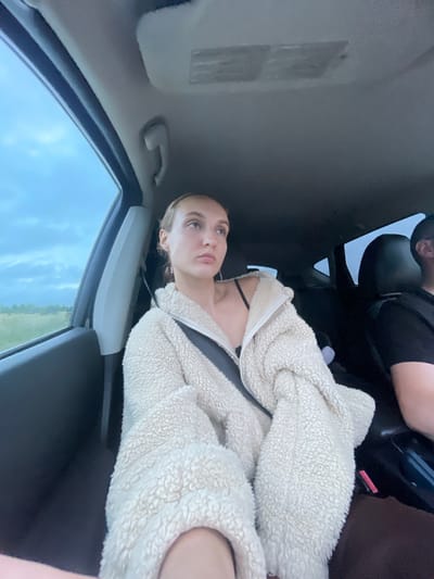 A medium shot captures a young woman with light brown hair pulled back, wearing a cream-colored fuzzy jacket and a black bra, seated in the passenger seat of a vehicle. Her gaze is directed to her right, with a thoughtful expression. A seatbelt is fastened across her chest. The interior of the car is visible, including the ceiling and part of the driver's seat and shoulder in the background. Through the car window, a cloudy, overcast sky and a flat, grassy landscape can be seen, suggesting a rural or roadside setting. The location is identified as Meshcheryaki, Russia.