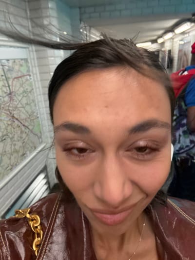 An inverted close-up selfie captures a person looking directly at the camera with a slight smile. The individual has dark hair pulled back, dark eyebrows, and brown eyes that appear to be made up with eyeshadow. A small, white earbud is visible in their right ear. They are wearing a delicate gold-colored chain necklace with a small pendant.

Part of a glossy, dark brown garment or bag with prominent gold chain details on its strap is visible on the right side of the frame. The background is mostly out of focus but reveals an indoor public transport setting, likely a metro or subway. Below the person's face, a blurry but discernible metro map, consistent with a Paris Metro map, is seen printed on a surface. Above and behind them, light-colored structures and linear lighting fixtures suggest an escalator or the interior of a train car or station. Other blurry figures, one wearing blue and another red, are faintly visible in the upper background. The overall mood conveyed by the person's expression is calm and pleasant. No specific text is clearly legible beyond the indistinct lines and colors of the map.