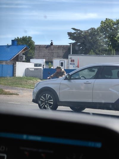 A man in a camouflage jacket and hat is riding a motorcycle down a paved road in Myadzyel, Belarus. He is in the middle of the frame, and the motorcycle is partially obscured by a silver car. The car is in the foreground, and the motorcycle is in the background. The man is looking forward, and the motorcycle is leaning slightly to the right. The road is wet and there are some trees and buildings in the background. The sky is blue and there are some clouds.