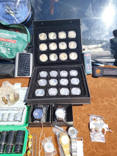 The image displays a collection of items laid out on a brown, textured surface, likely a market stall or a personal collection being sold. The setting appears to be outdoors, with bright sunlight casting shadows and creating reflections on the glass behind some of the objects. The time of day is clearly daytime, indicated by the strong sunlight.
The main subjects are various objects, including: a black velvet-lined case filled with what appear to be commemorative coins, both gold and silver-toned, each depicting faces or emblems; a metal flask with a textured, dark cover; a green bag with the "SPARK LUX" logo and text indicating "professional tools" and "power cables" in Russian; a box of socket wrenches; and several wristwatches, including a dark-faced analog watch and a skeletonized silver watch. There are also some pieces of jewelry, including beaded necklaces and what look like earrings or brooches presented on a card. A small box with a red star emblem and the word "NATO" is also visible, as is a small red pouch with a yellow star.
The overall impression is of a display of goods for sale, possibly at a flea market or outdoor bazaar. There are no people visible in the frame. The condition of the items suggests they are either vintage, collectible, or new merchandise being offered. The presence of Russian text on the "SPARK LUX" bag and other items suggests the location is likely in Russia, confirmed by the provided location context of Chesnokovka, Russia. The scene is illuminated by natural daylight, creating a bright and somewhat harsh lighting effect.