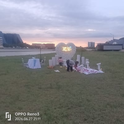 The image depicts an outdoor proposal setup on a grassy area next to a body of water, likely a river or sea, with buildings visible on the opposite bank. The scene is set during dusk, with a soft, hazy sky illuminated by the fading sunlight.
The main focus of the setup is a large, heart-shaped balloon arch adorned with numerous white flowers. A neon sign within the heart reads, "Will You Marry Me?". Around the heart, several tall white cylindrical pillars and white vases filled with white flowers are strategically placed. A white cloth is spread on the ground, decorated with scattered red rose petals and lit candles, creating a romantic ambiance.
Two people are present. One individual, wearing dark pants and a gray shirt, is actively arranging the rose petals. Another person, a child in a pink jacket, is standing near the balloon heart. A small dining table set for two, draped with a white tablecloth and a light blue runner, is visible to the left of the heart, suggesting a romantic dinner setting.
In the background, a modern, multi-story building with a distinctive tiered design overlooks the water. Other buildings and structures are also visible in the distance, along with streetlights. The overall mood is one of anticipation and romance, as the scene is meticulously prepared for a marriage proposal.
The bottom left corner of the image contains text that reads "OPPO Reno3" and "2026.06.27 21:11", indicating the device and date/time the photo was taken.