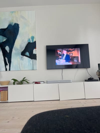 The image depicts an indoor living space, identified as a residence in Skanderborg, Denmark. A large, wall-mounted black flat-screen television is centrally positioned on a white wall. Cables are visible extending downwards from the television. To the left, a tall, rectangular abstract painting features dark blue and black shapes on a lighter blue, cream, and white background, with visible paint drips.
Below the television, a white, modular media cabinet composed of multiple closed compartments spans the wall. A silver-grey Sonos soundbar rests on the cabinet directly beneath the television. Various objects are arrayed on the cabinet: a stack of books and a light-colored wooden bowl are on the far left. To their right, a green-leafed plant is contained within a clear glass bowl. Further right, a small red model sports car and a dark rectangular object are present. On the far right, a black gaming controller and a portion of a metallic, possibly electronic, device are visible.
The television screen displays an interior scene. A man dressed in a suit is seated at a table, raising a glass. An arm is partially visible to his left. In the background, a large, dark silhouette of a fish (resembling a marlin or swordfish) is mounted on a wall, suggesting a restaurant or themed establishment. The floor consists of light-colored wooden planks. The lower portion of the foreground is occupied by a dark, textured surface, likely part of a sofa or chair from which the image was captured.