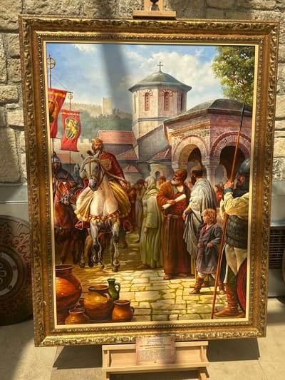 This is a detailed oil painting displayed in a museum, likely in the city of Neofit Rilski, Bulgaria, as suggested by a label at the bottom of the frame. The painting depicts a historical scene with a prominent figure on horseback at its center.

The central subject is a regal man wearing a golden crown and an ornate red and gold robe, seated on a white horse. The horse has a decorative bridle and saddle cloth. To the left of the central figure, another man on horseback, dressed in armor, follows closely. Two large red banners with gold emblems are displayed on poles to the left, suggesting a royal procession or arrival.

In front of the horses, a group of people are gathered. Some are dressed in tunics and robes of various colors, including red, brown, and cream. One man in a brown robe holds a rolled scroll. A young boy in a gray tunic stands among the crowd. Behind this group, there are buildings with arched doorways and red-tiled roofs, characteristic of historical architecture, possibly a church or monastery with a dome topped with a cross. A larger fortress wall is visible in the background under a blue sky with scattered clouds.

To the right of the main group, a man in armor holding a spear stands guard. A large shield with a decorative pattern is leaning against a wall on the far left. In the foreground, several clay pots and a green jug are arranged on a stone-paved ground. The overall scene conveys a sense of historical importance and ceremony. A wooden stand with an informational plaque is placed in front of the painting.
