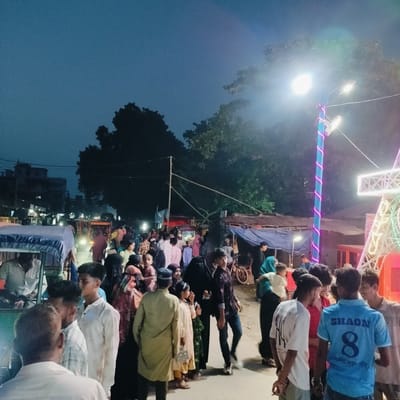 This vibrant image captures a bustling evening or night scene on a crowded street in Charfasson, Bangladesh. The dark blue sky indicates dusk or nighttime, with the area brightly illuminated by numerous artificial lights, including streetlights and decorative installations.

The scene is densely populated with a diverse group of people of various ages, including men, women, and children, many appearing to be socializing, walking, or observing their surroundings. Their attire is casual and typical of the region.

Prominent objects in the image include a partial view of an auto-rickshaw with a blue canopy on the left. Several bright streetlights are visible, with one on the right creating significant glare. The pole holding this light is adorned with spiraling pink and purple decorative lights. To the right, a large, intricate white structure, possibly part of a fairground ride or a decorated gate for an event, is illuminated with internal colorful lights. In the background, obscured by the crowd and darkness, are simple buildings and market stalls covered with blue tarps and corrugated iron, suggesting a temporary market or festive gathering. Overhead, power lines crisscross the sky.

The overall atmosphere appears lively and festive, consistent with a community event or an evening market. The decorative lights strongly suggest a celebration or festival is taking place, potentially Eid, given the visible text.

On the back of a blue T-shirt worn by a young man in the foreground, the name "SHAON" and the large number "8" are clearly visible in white text. Below the number, Bengali script can be discerned, which reads "শুভ ঈদ মোবারক" (Shubho Eid Mubarak), directly indicating an Eid celebration.