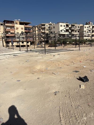 The image displays an unpaved urban lot in Mu'adammiyat ash-Sham, Syria, under a clear blue sky. The foreground consists of bare, sandy ground strewn with scattered debris, including a prominent black plastic bag, a small light-colored book or box, and miscellaneous litter. Tire tracks are visible across the dirt. A human shadow, likely that of the photographer holding a device, appears in the lower left corner.
In the midground, a grid of black metal frameworks, appearing as incomplete or disassembled market stalls, occupies a significant area. These structures comprise multiple vertical posts connected by horizontal beams; some beams lie on the ground alongside the upright sections.
The background features a dense block of multi-story residential buildings constructed from concrete or stone, exhibiting various states of finish. Visible architectural details include balconies, rooftop satellite dishes, and water tanks. Laundry is seen hanging from a balcony on the leftmost building. Sparse green foliage and one or two parked vehicles are discernible between the metal frames and the residential structures. No individuals are actively present or engaged in the visible scene apart from the photographer's shadow.