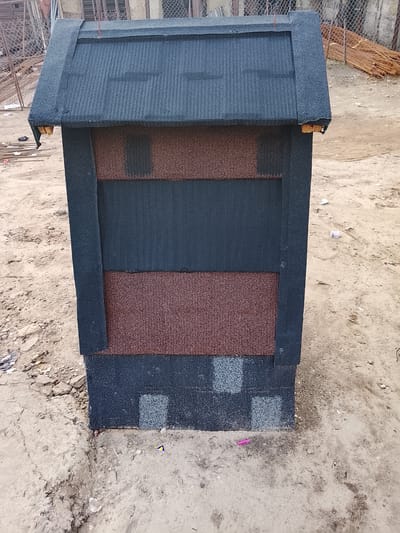 A dark gray, gable-roofed structure, likely a small house or shed, stands on a dirt ground in Rumuepirikom, Nigeria. The roof is covered with dark gray shingles. The walls of the structure are a combination of dark gray and reddish-brown panels, also made of shingles. The bottom portion of the walls features a pattern of square, light gray shingles. The background shows a chain-link fence, some rebar, and scattered debris. The ground is a mixture of sand, dirt, and small stones. There are no people visible in the image.