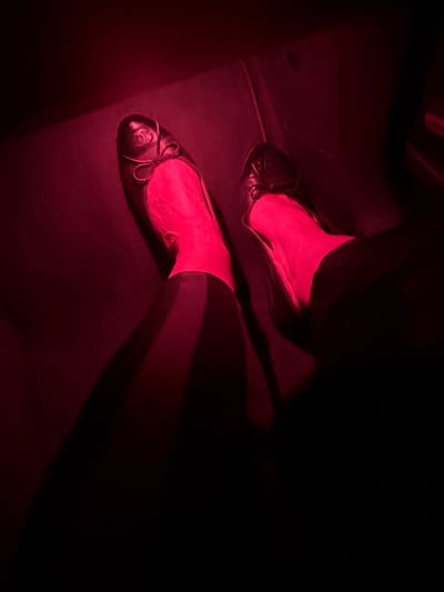 The image presents a downward-angled view of a person's lower legs and feet, situated within a dimly lit interior primarily illuminated by a strong red light source. The individual wears dark hosiery or tights and two black ballet flats. The left shoe features a prominent Chanel "CC" logo on its toe, accompanied by a small black bow. The right shoe also has a small black bow on its toe. Both feet rest on a dark, flat surface, potentially a vehicle's footwell, which is also bathed in the red light. Elongated shadows from the legs and feet are cast onto this surface. No specific location within Vagos, Portugal, is discernible from the visual information provided.