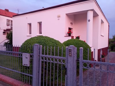 The image shows a modern house with white walls and a dark brown roof. The house has several windows and a prominent column supporting an overhang. A number "27" is visible on the exterior wall. In front of the house, there are two large, rounded green bushes. A grey metal fence with a decorative circular pattern runs along the front of the property. A mailbox is attached to the fence. Red flowers in pots are visible near the entrance of the house and to the right of the house, one in a wooden barrel. The ground in front of the house is paved with grey stones. The sky is overcast and appears to be dusk. The scene is located in Kołobrzeg, Poland, as indicated by the metadata. There are no people visible in the image.