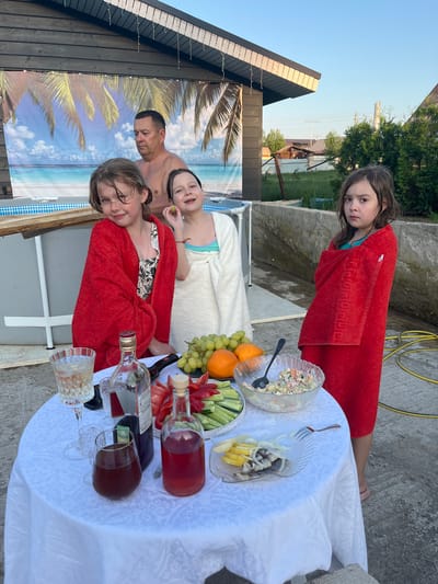 A group of people are gathered near a pool, likely for a meal after swimming. The scene is outdoors, possibly a backyard in Новая Чернушка, Russia. Three girls are present, two wearing red towels and one in a white towel. A man with no shirt is also present. A table covered with a white tablecloth holds a variety of food and drinks, including salad, fruit, vegetables, and bottles of liquid, possibly wine. There are also glasses. A backdrop with a beach scene is set up behind the pool.