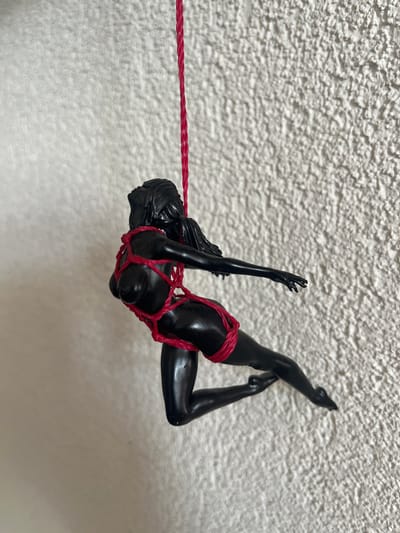 This media file features a black, glossy figurine of a woman suspended by a red rope against a textured, white background. The figurine is posed in a dynamic, contorted position, with its limbs outstretched and its body bound by the rope in a way that resembles bondage. The rope extends horizontally across the frame, tethering the figurine from its torso. The texture of the background suggests a wall or ceiling with a rough, stucco-like finish. The lighting is bright and appears to be coming from the right, casting subtle shadows that highlight the glossy surface of the figurine and the texture of the background. There are no discernible location cues, time of day, weather, or visible text in the image. The overall impression is one of artistic presentation, possibly a sculpture or a decorative item.