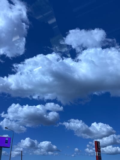 The image is a low-angle shot looking up at a bright blue sky filled with fluffy white and gray clouds. A bright, horizontal band of light, possibly from a plane contrail or a lens flare, is visible within a large cloud in the upper center of the frame. In the lower left corner, a lamppost extends upwards, and to its left, a billboard with a purple background displays an image of a bottle. In the lower right, a tall red sign with white lettering and a logo is visible. The environment suggests an outdoor urban setting, possibly near a road, given the presence of the billboard and lamppost. The image appears to be taken from a moving vehicle, as evidenced by reflections on the glass surface at the top of the frame. The location is identified as Poroshkino, Russia.