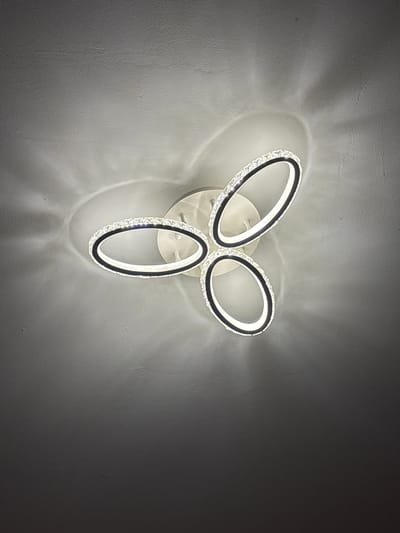 A modern, three-ringed pendant light fixture with integrated LED lighting is attached to a textured ceiling. The fixture is white and features oval-shaped rings adorned with clear crystals and black trim. The lighting casts a diffused glow and shadow patterns onto the ceiling. The image is a close-up shot, focusing on the light fixture and the surrounding ceiling texture. There are no people or other objects visible in the frame. The location is identified as Marchevo, Bulgaria.