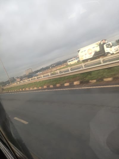 The scene depicts a multi-lane highway under an overcast sky, located in Murera ward, Kenya. The foreground shows an asphalt roadway with a faded yellow lane marking in the bottom-left. A central median separates traffic lanes, consisting of green grass bordered by a concrete curb painted with alternating black and white/yellow sections. Metal guardrails run along the far side of the median. Vehicles are visible on the distant lanes, including a white box truck with "Maziwa freshi" branding and a red-orange logo, moving rightward. Behind it and to its left is a white passenger van. Additional indistinct vehicles are also present on these lanes. In the background, a skyline with multiple-story buildings and distant vegetation is visible. The image exhibits reflections on glass in the bottom left, consistent with being taken from inside a moving vehicle. No individuals are discernible within the frame.