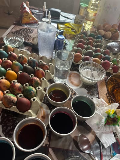 A table is covered with items related to egg dyeing, including two egg cartons, one containing undyed eggs and another with dyed eggs. There are cups filled with various colored dyes, a glass bowl with white powder (likely for making the dye mixture), a glass of water, paper towels with dye stains, paintbrushes, and a spoon. A jar of oil and other objects are present on the left side of the image. The background includes a patterned table cloth that may be representing coffee beans. The location is identified as Galiche, Bulgaria, based on associated information. No people are visible in the scene.
