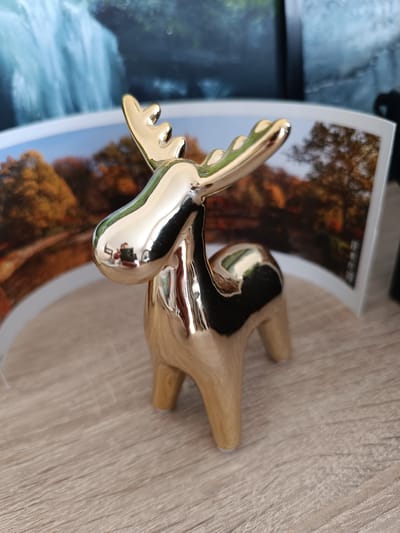 A small, shiny, gold-colored ceramic figurine of a moose stands on a light wood-grain surface. The moose has antlers, a rounded body, and four legs. The figurine is highly reflective, mirroring the surroundings. In the background, a curved photograph or print is visible, depicting a scene with autumn trees and a body of water, and a large waterfall. A person's reflection, holding a smartphone, is visible on the moose's head. The lighting suggests it is daytime. The location context is Olsztyn, Poland.