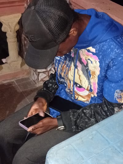In Ukpor, Nigeria, a person wearing a black baseball cap and a blue hoodie with a graphic print is seated, looking down at two mobile phones. One phone, purple, is held in their hands, while another, black, is placed on their lap. The person is wearing dark gray pants and a black, patterned jacket. The environment appears to be outdoors, with a decorative pillar and tiled flooring visible. A light blue plastic table is also present in the foreground.