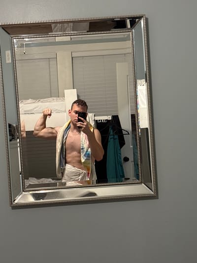A young adult male is standing in front of a mirror, taking a selfie. He is flexing his bicep and wearing a towel around his waist. A colorful towel is draped over his shoulders. The mirror reflects a room with bunk beds and clothing hanging on a rack. The image is taken indoors in what appears to be a bedroom or dorm room. The walls are painted a neutral gray color. The mirror has a decorative silver frame. The location is Panama City Beach, United States of America.