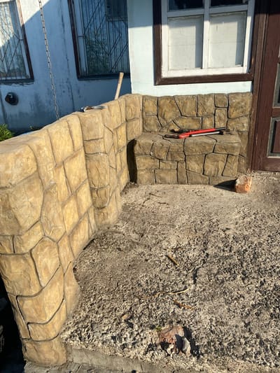 This media file depicts an outdoor scene in Vasylky, Ukraine, likely in front of a building. The primary subject is a set of stairs and a small landing constructed with stamped concrete that imitates the appearance of natural stone blocks. The concrete has a textured, tan and brown finish.
To the left of the stairs, a section of a building is visible. It features a window with white trim and a dark wooden frame, divided into panes. Below this window, another window with a metal grate is partially visible, and a chain hangs across the building's facade. The building's exterior wall appears to be painted a light blue, with areas of peeling paint and what looks like moss or dirt accumulation.
On the stone-patterned landing, near the top of the stairs, a pair of red and black bolt cutters are leaning against the wall. The wall itself is constructed with large, irregular stone-like shapes, also made of stamped concrete. To the right of the stairs and landing, a rough, textured wall, possibly made of concrete or a similar material with visible aggregate, rises steeply. Scattered on this wall and the ground are small branches and debris, including a fragment of red brick.
The lighting suggests it is daytime, with clear sunlight casting shadows, indicating fair weather. There is no visible text in the image. The scene appears to be part of an exterior renovation or construction project, given the presence of the construction tool and the textured, unfinished look of the wall to the right. The overall impression is of an unfinished or rustic exterior entrance.