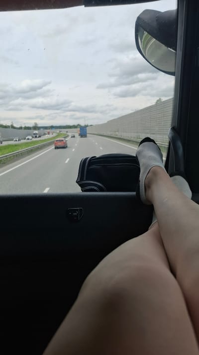 The image shows a person's legs and feet, wearing white socks with black trim, resting on a dark surface inside a vehicle. The person's legs are crossed, and their skin appears fair with some minor marks. They are positioned to look out the window, where a highway scene is visible.
The scene outside the vehicle is a multi-lane highway with other vehicles, including cars and a truck, traveling in the same direction. The highway is bordered by green grass and a concrete barrier. The sky is cloudy, suggesting overcast or partly cloudy weather. The perspective suggests the photo was taken from the passenger seat of a vehicle. There is a side-view mirror visible on the left side of the frame, reflecting the sky and a distant green area. On the dark surface in the foreground, there is a small black object with white text that reads "SOS" with a small asterisk.
The overall impression is a moment of travel, possibly on a road trip, with the focus on the relaxed posture of the passenger. The lighting indicates it is daytime. The specific mention of "Kurmanka, Russia" provides a geographical context for the location.
