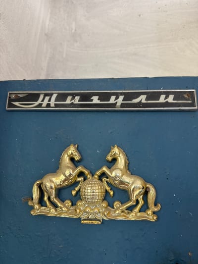 An ornate brass emblem featuring two rearing horses on either side of a globe rests on a dark blue surface. Above the horses, a silver metallic emblem with dark lettering, possibly a car badge, is affixed to a lighter, textured surface. The lettering on the silver emblem appears to be in Cyrillic script. The scene is indoors, with a plain, light-colored wall above the emblems. The overall impression is that of a decorative element on a vehicle or a building facade. The provided context of Khazar Raion, Azerbaijan, suggests a potential location for this emblem, perhaps on a historical vehicle or a building associated with the region.